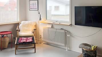 Un salon comprenant un fauteuil, une petite table, un radiateur, un téléviseur et une fenêtre donnant sur des immeubles.