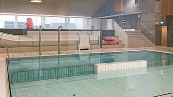 Piscina interior con suelo de baldosas, una escalera que lleva a una zona de sauna y vistas al exterior a través de amplias ventanas.
