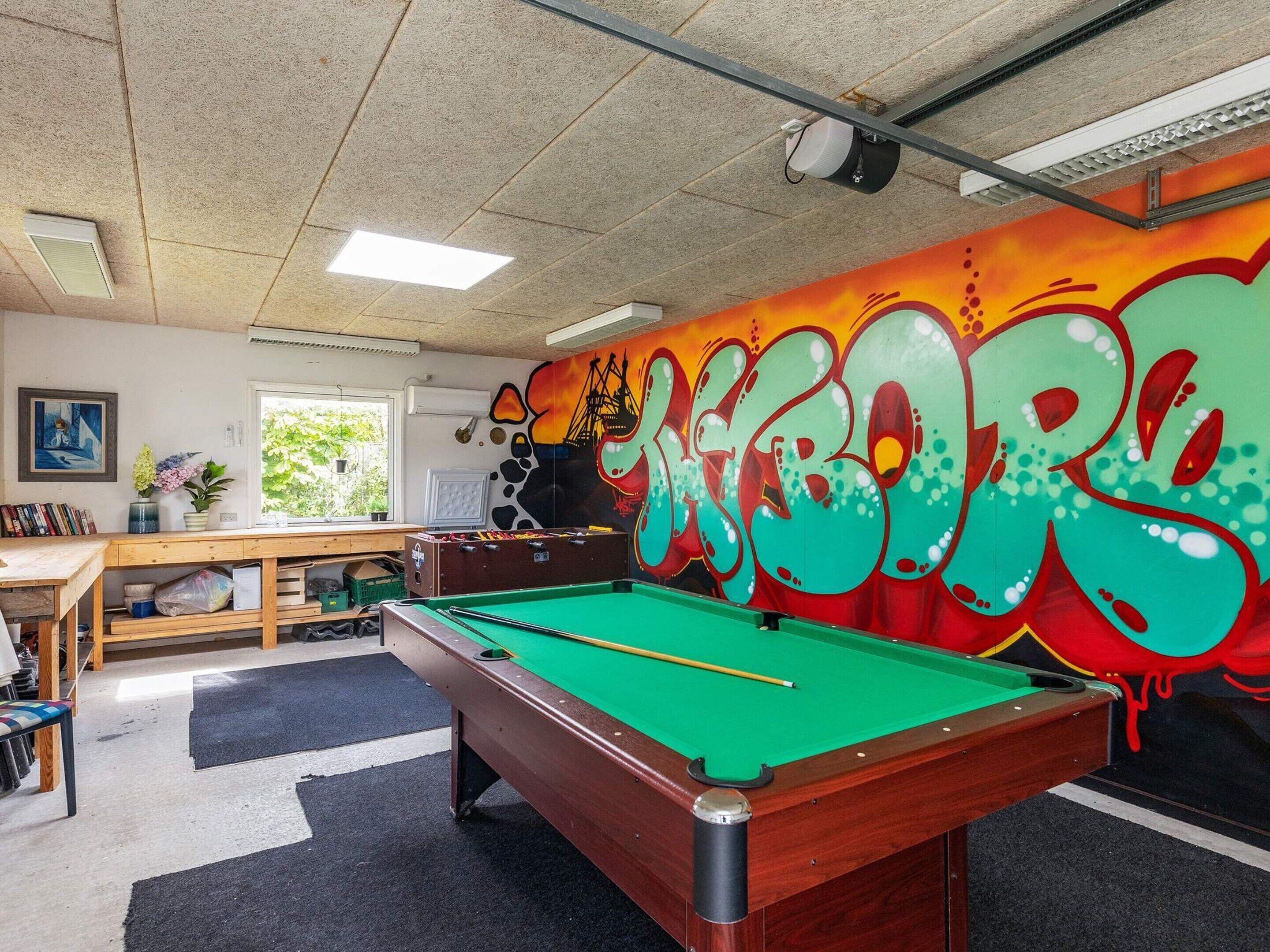 Ein Billardtisch mit grüner Spielvliesdecke, ein Holztisch mit einem Laptop und eine farbenfrohe Graffiti-Wand.