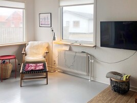 A living room with a chair, a small table, a radiator, a TV, and a window with a view of a house.