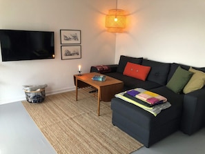 A living room with a flat-screen TV, a coffee table, a sofa with cushions, a wall-mounted lamp, and a small basket. - 6 Person Holiday Home in Thyboron-by Traum (Thyborøn)