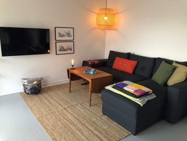 A living room with a flat-screen TV, a coffee table, a sofa with cushions, a wall-mounted lamp, and a small basket. - 4 Star Holiday Home in Thyboron (Thyborøn)