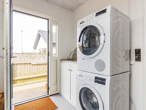A compact laundry room with a stacked washer and dryer, a small countertop, and a sliding glass door leading to an outdoor area. - 4 Star Holiday Home in Thyboron (Thyborøn)