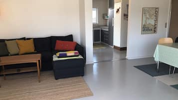 Una sala de estar con un sofá, una mesa de centro y un pequeño reposapiés con una manta colorida. Hay una mesa de comedor con mantel verde y una silla. Se ve una cocina al fondo.