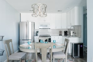 A modern kitchen with white cabinets, a stainless steel refrigerator, a microwave, and a sink. There is a dining table with chairs and a chandelier above.