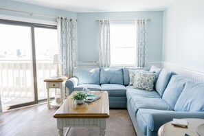 A room with a light blue sofa, a wooden coffee table with a plant, and a view of the outside through a large window.