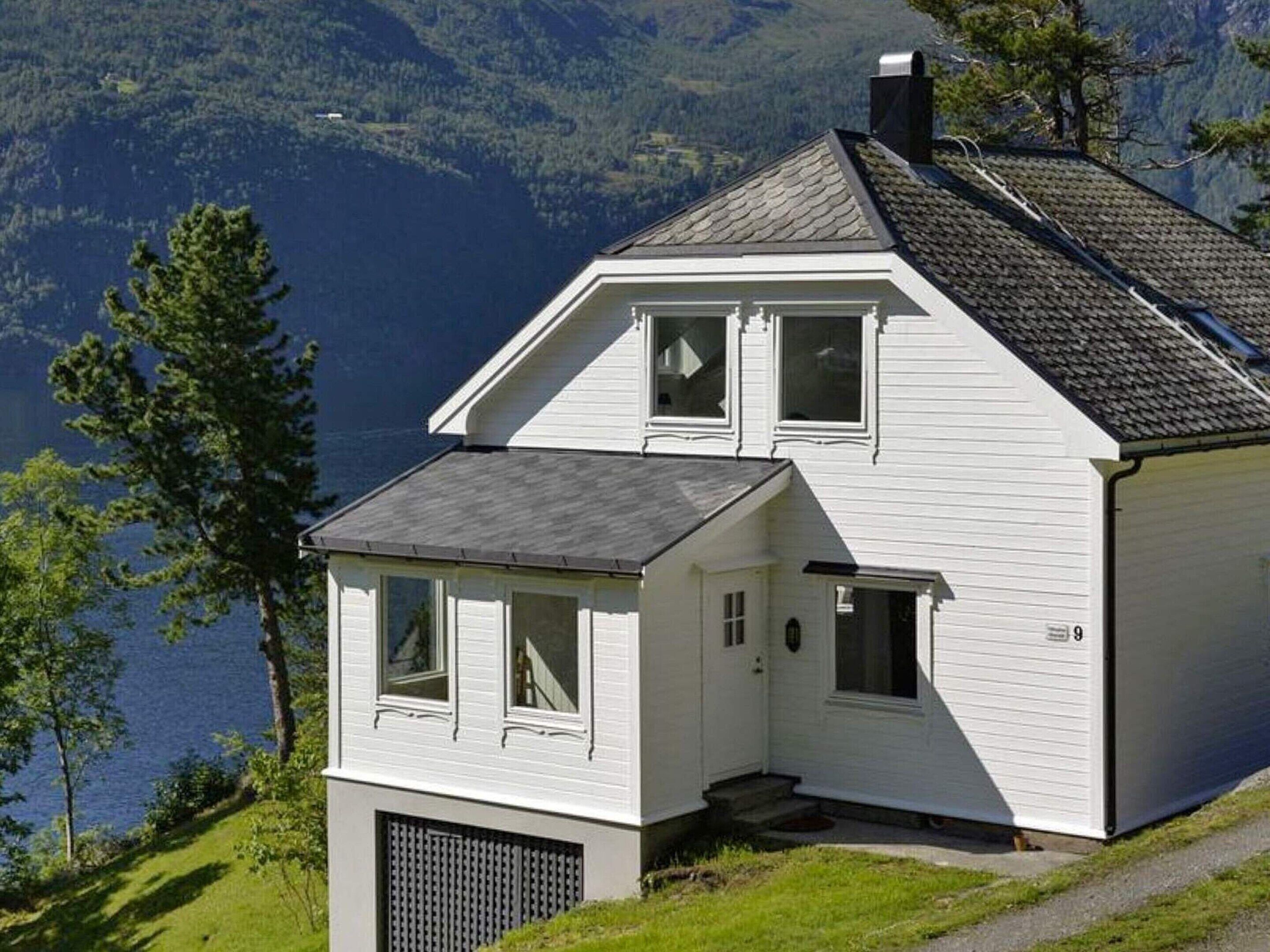 Een wit huis met twee verdiepingen, een schuin dak, een veranda en een garage. Het staat op een helling met uitzicht op een meer en beboste bergen.