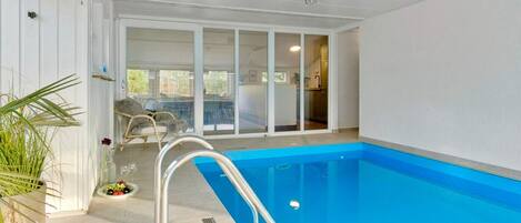 An indoor swimming pool with a clear view of the adjacent room, a chair, and a potted plant.