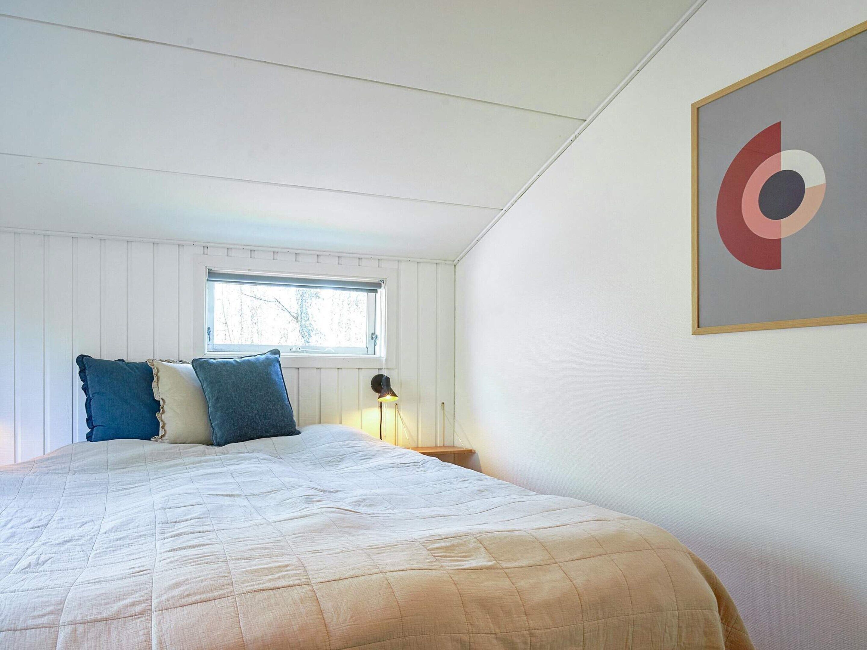 Un dormitorio con cama, almohadas, mesita de noche, lámpara, ventana y una obra de arte abstracto enmarcada en la pared.