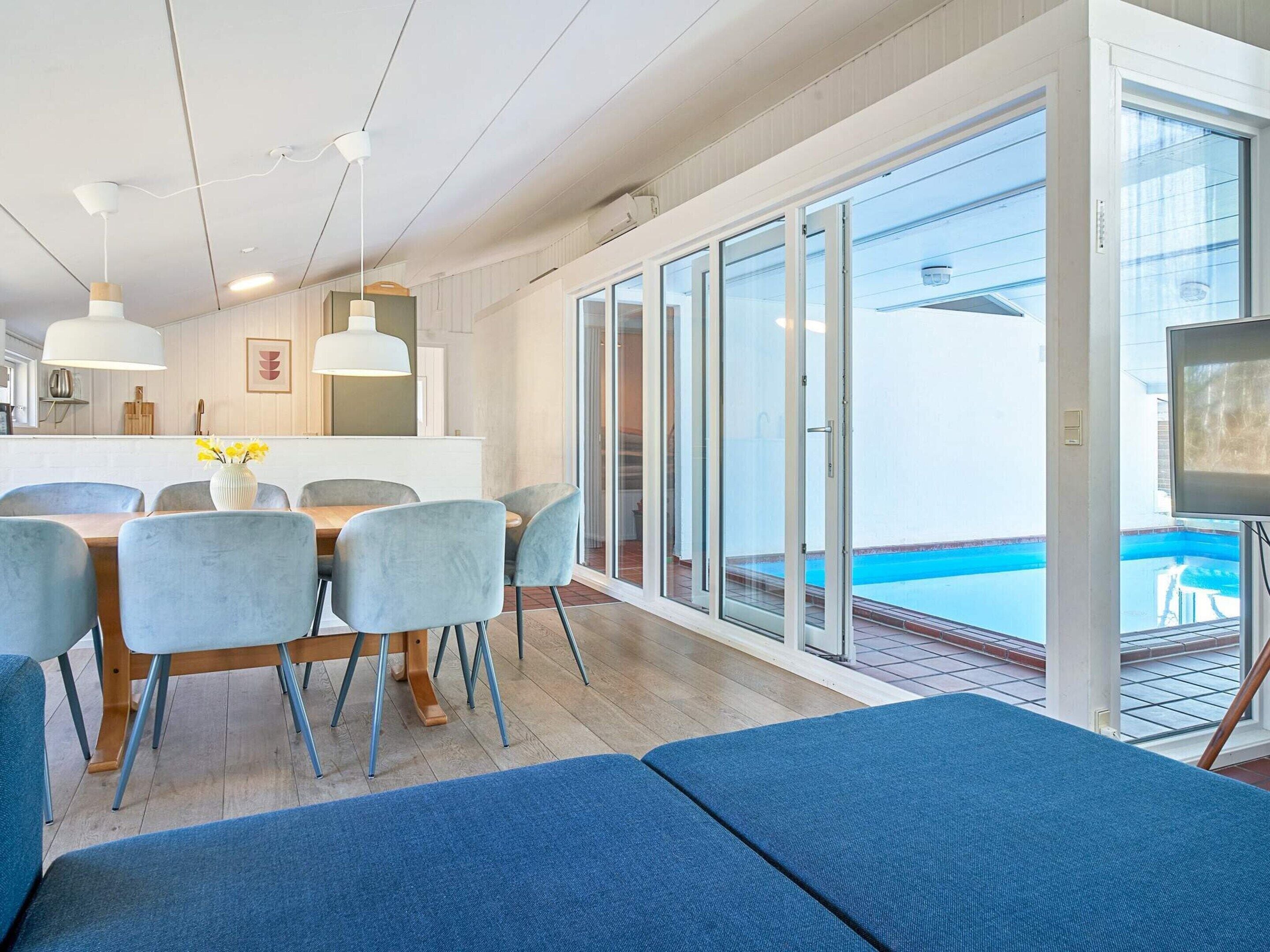 A modern dining area with a pool view, featuring a table with chairs, a sofa, and a TV.