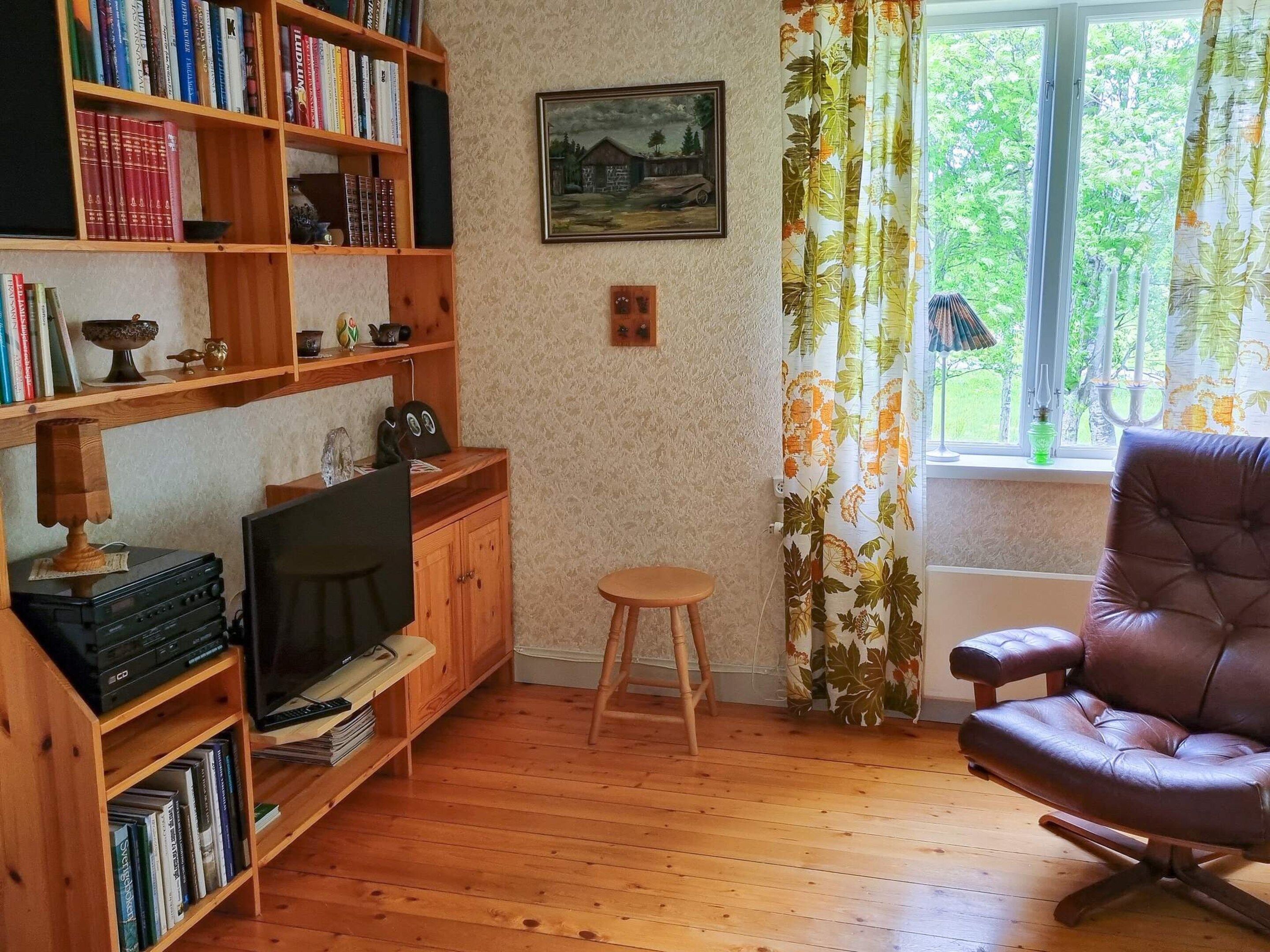 A cozy room with a wooden floor, a leather chair, a television, a bookshelf, and a window with curtains.
