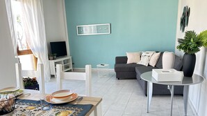 A living room with a sofa, a small table, a TV, and a dining area with a table set for two. - Lara's Apartment Mare (Lido di  Camaiore)