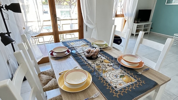 A dining area with a table set for four, white chairs, a woven chair cushion, a TV stand, and a view of the outdoors. - Lara's Apartment Mare (Lido di  Camaiore)