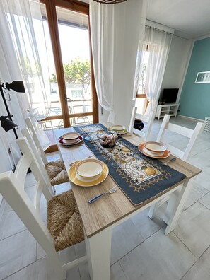 A dining area with a table set for four, white chairs, a woven chair cushion, a TV stand, and a view of the outdoors. - Lara's Apartment Mare (Lido di  Camaiore)