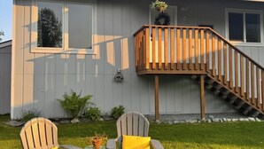 A two-story house with a wooden staircase, two Adirondack chairs with yellow cushions, and a small garden area.