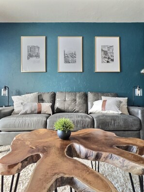 A living room with a grey sectional sofa, three framed black and white photographs on the wall, a small potted plant on a wooden coffee table, and two floor lamps on either side of the sofa.