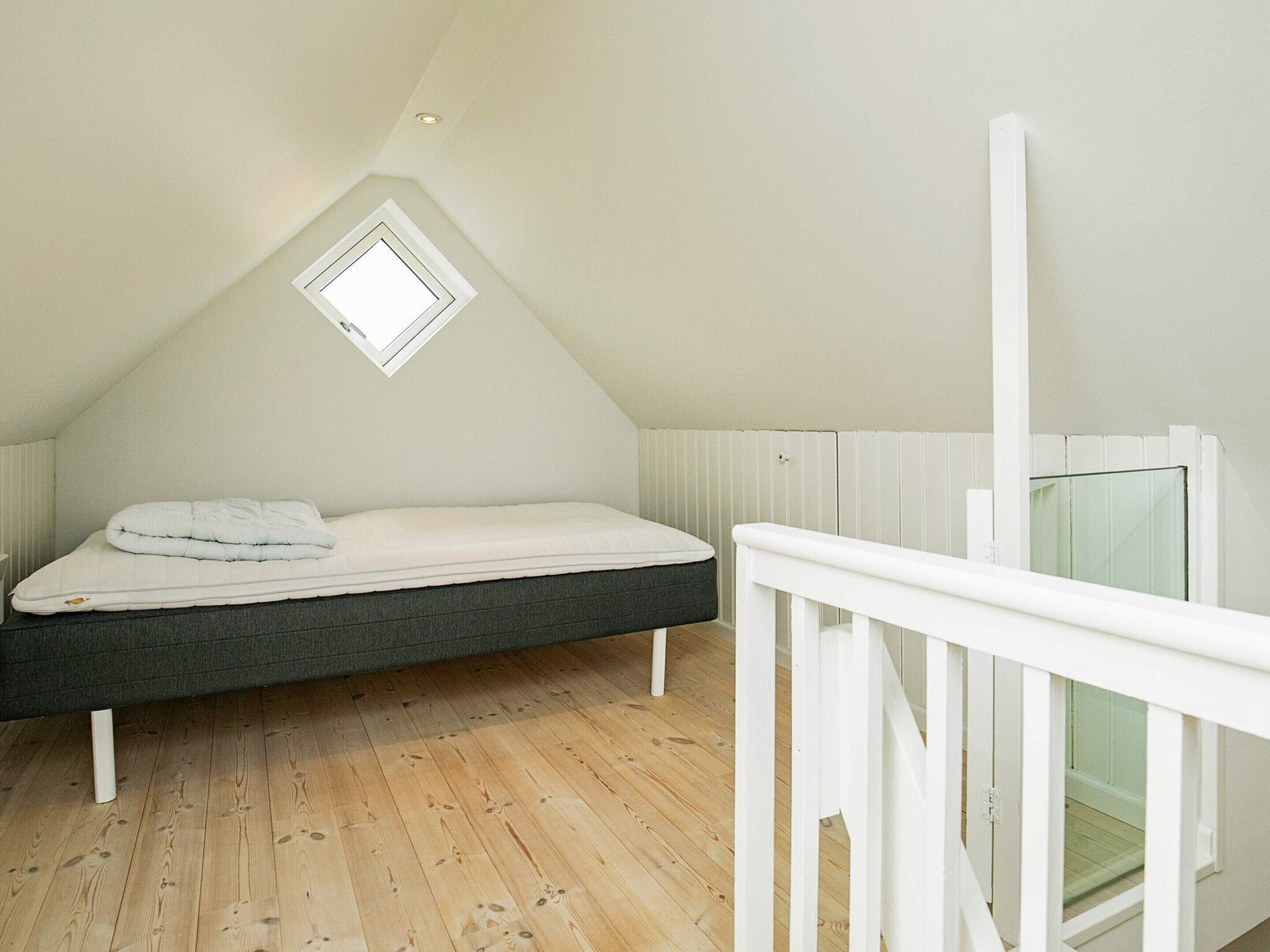 Une chambre avec un plafond en pente, un lit simple, une petite fenêtre et un plancher en bois.