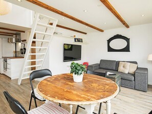 A modern living room with a round wooden table, a grey sofa, a TV mounted on the wall, and a white ladder leading to an upper level.