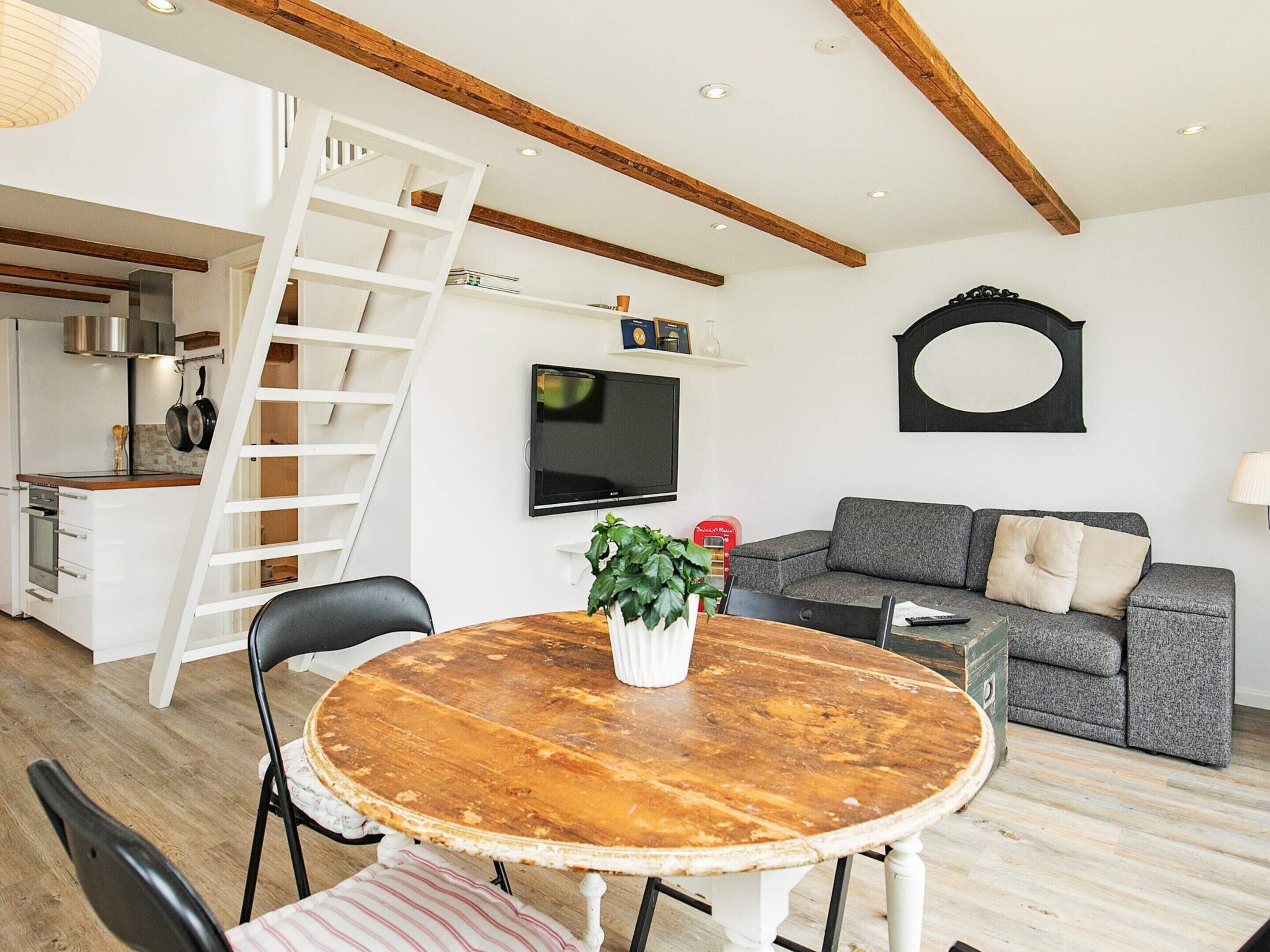 A modern living room with a wooden dining table, a gray sofa, a TV mounted on the wall, and a wooden ceiling with exposed beams.