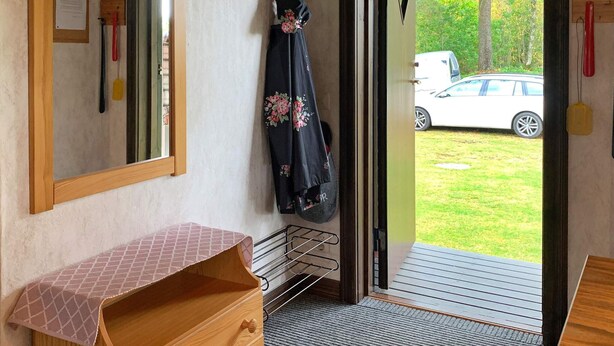 Une pièce avec une commode en bois, un miroir, un porte-manteau sur lequel est accroché un vêtement à motifs floraux, et une vue sur une voiture blanche par une porte ouverte.
