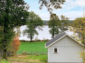A small, single-story cabin with a gabled roof, situated in a grassy field with a lake in the background.