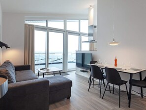 A modern living room with a sofa, a dining table with chairs, a flat-screen TV, and large windows offering a view of the sea.