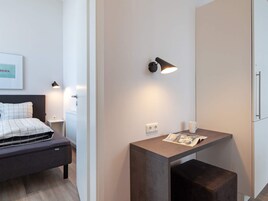 Una habitación de hotel moderna con cama, mesita de noche, lámpara y un pequeño escritorio con una revista.