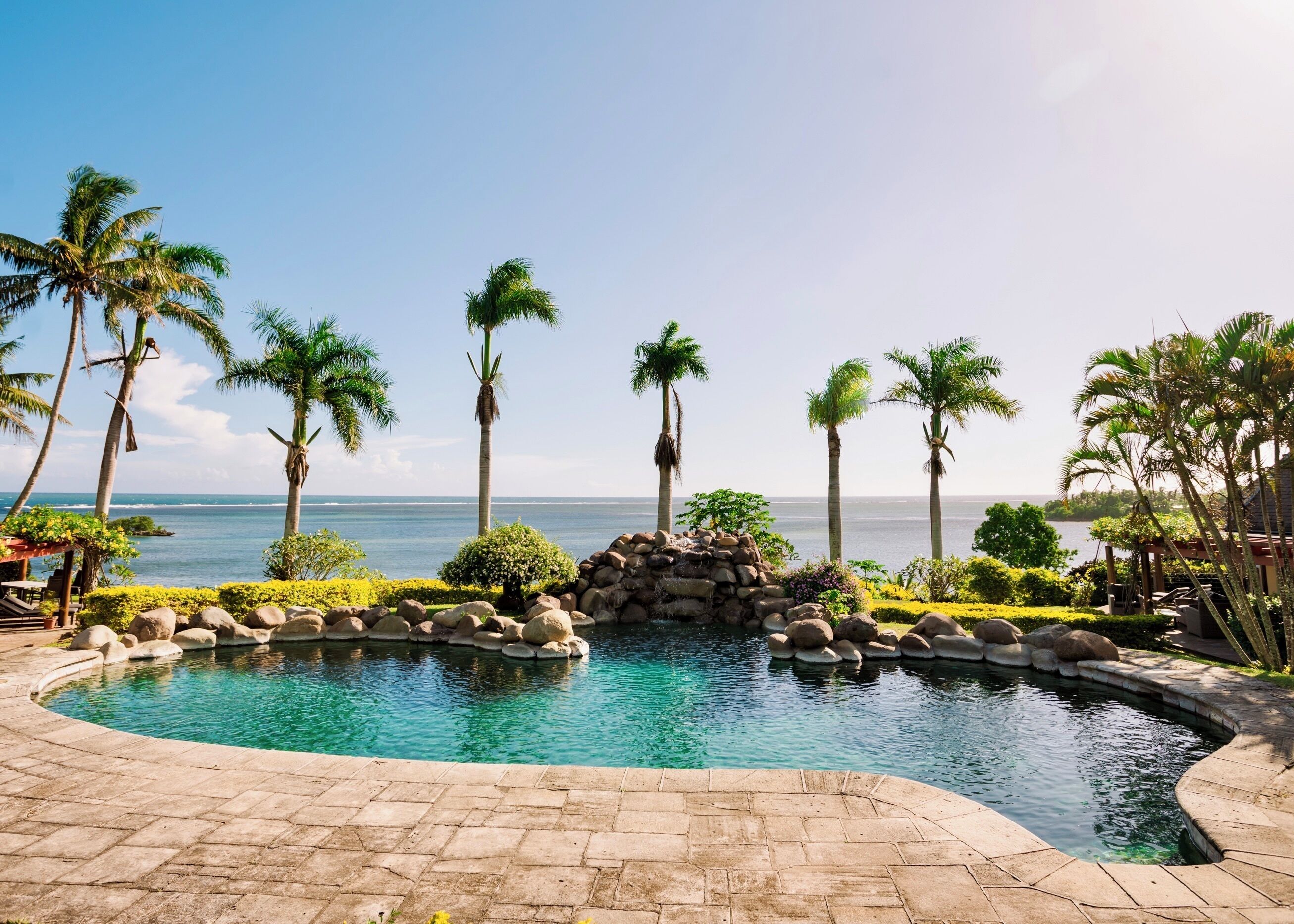 A pool with a view of the ocean, surrounded by palm trees and a stone wall.