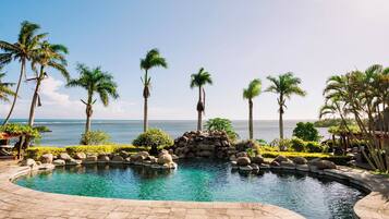 A pool with a view of the ocean, surrounded by palm trees and a stone wall.