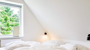 Ett sovrum med ett sluttande tak, en säng med vita sängkläder, ett fönster med utsikt över grönområden och en vägglampa.
