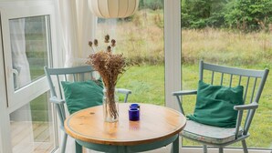 A round wooden table with a vase of dried flowers and a blue mug, flanked by two chairs with green cushions, set on a balcony with a view of a grassy area and trees. - 6 Person Holiday Home in Lokken (Lokken)