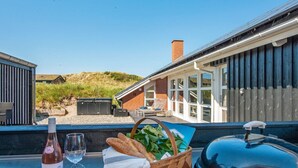 Un tavolo con una bottiglia di vino, bicchieri, pane e un cesto di verdure, con vista su una casa moderna con ampie finestre e sulla spiaggia.