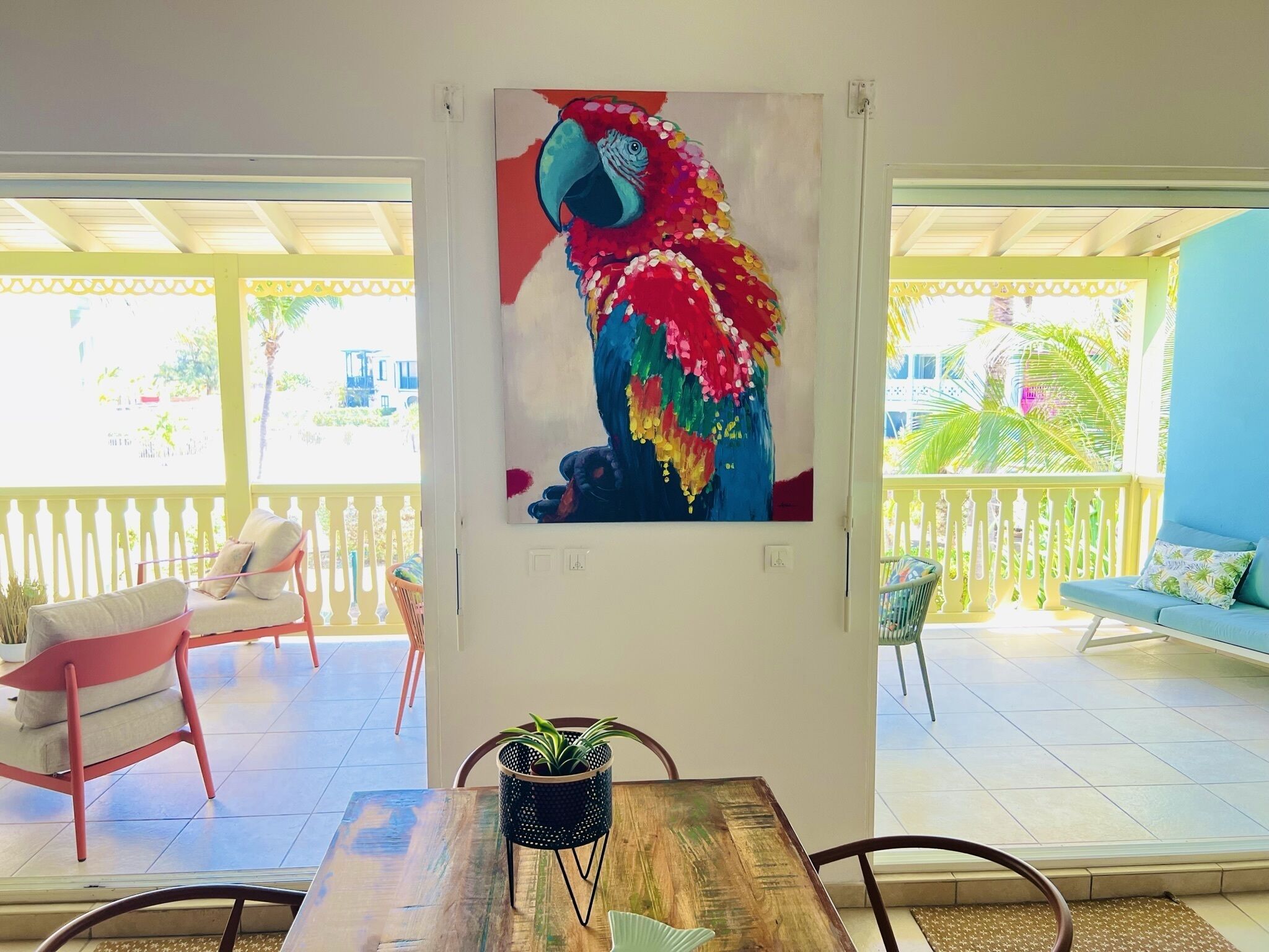A room with a wooden dining table, a chair, a sofa, and a colorful painting of a parrot on the wall.