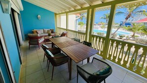 A covered patio with a wooden table and chairs, a sofa, and a view of a pool and palm trees.