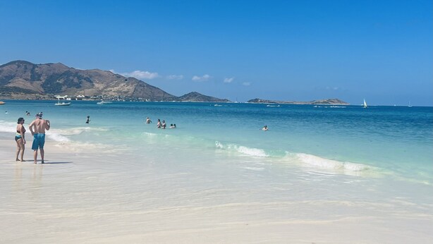 A beach with clear turquoise water, people swimming and sunbathing, and boats in the distance.