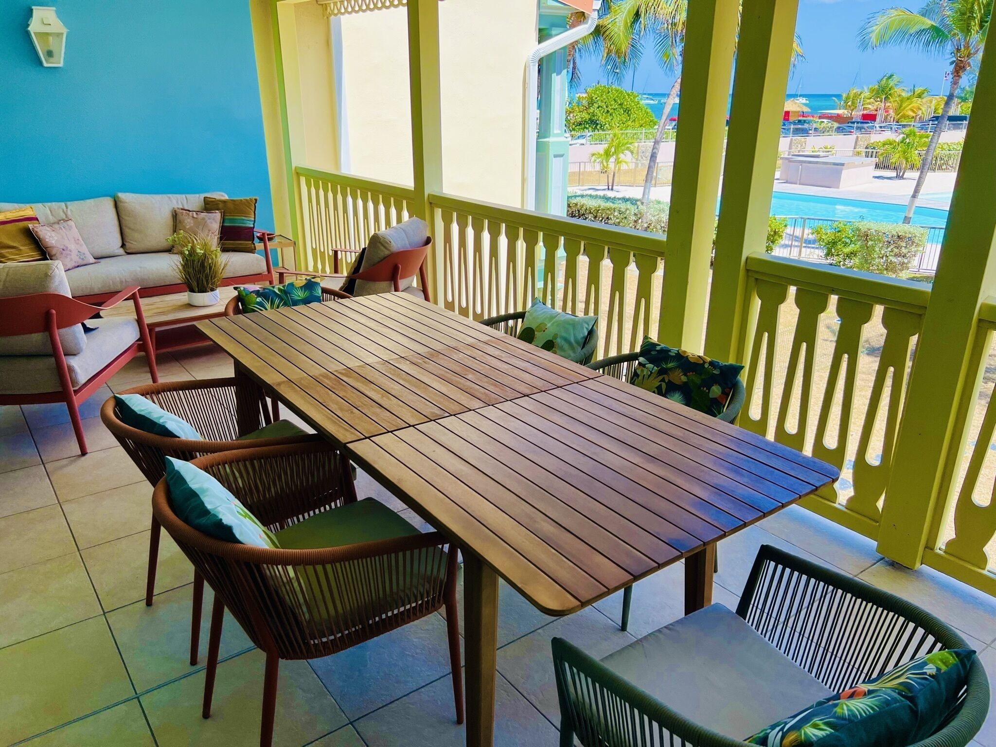 A balcony with a wooden table and chairs, a sofa, and a view of the ocean and palm trees.