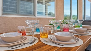 Mesa de comedor servida con varios cuencos, platos y vasos, con vista al exterior a través de grandes ventanales.