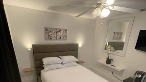 Une chambre moderne avec un grand lit, un ventilateur de plafond, une table de chevet avec une lampe et un petit bureau avec une chaise.