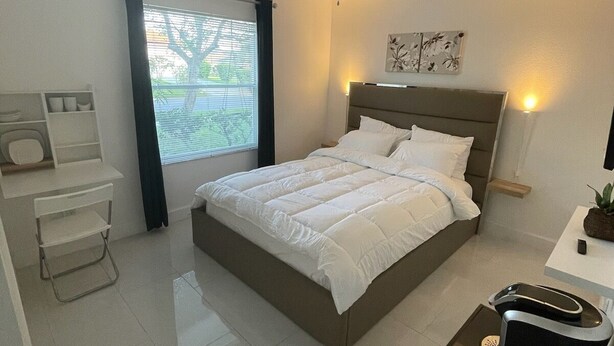 Une chambre moderne comprenant un lit, un bureau, une chaise, un ventilateur de plafond et une fenêtre avec des stores.