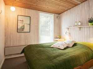 A bedroom with a wooden ceiling, a bed with a green quilt, a window with blinds, a wall-mounted picture, and a shelf with decorative items. - 6 Person Holiday Home in Blavand (Blavand)