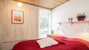 A bedroom with a red quilted bedspread, a white pillow, a wooden ceiling, a window with blinds, and a wall-mounted shelf with plants. - 6 Person Holiday Home in Blavand (Blavand)