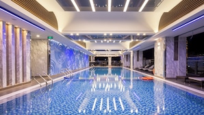 Uma piscina coberta espaçosa com chão de azulejos quadriculados, rodeada por paredes com acabamento em mármore e lambris de madeira.