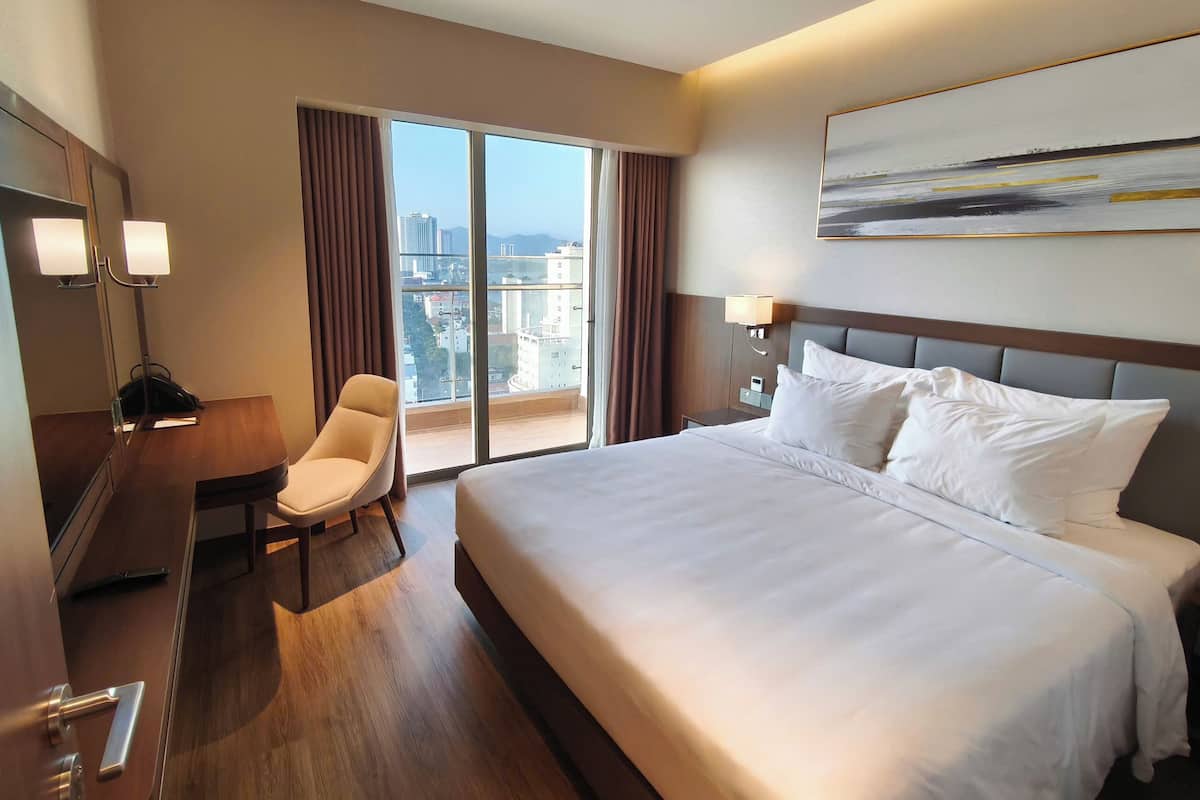 Habitación de hotel con una cama grande, un escritorio, una silla y vistas de la ciudad a través de la ventana.