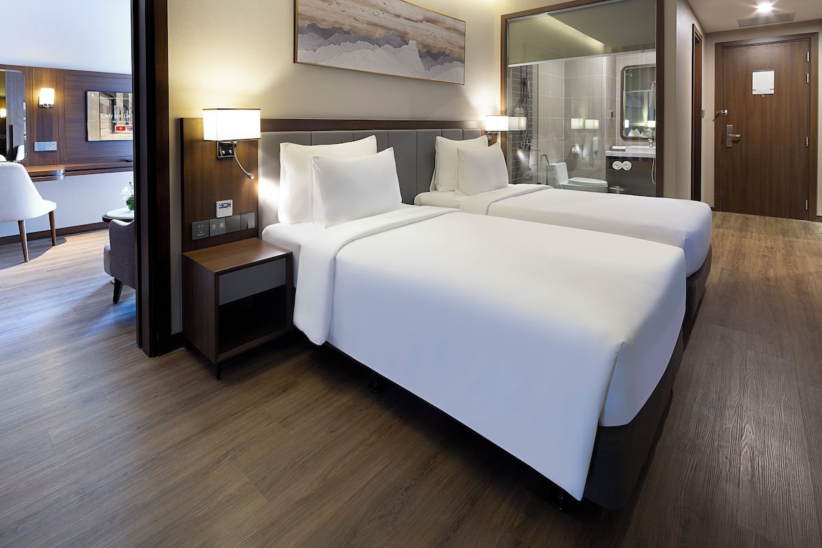Habitación de hotel con dos camas, una mesita de noche, un escritorio y un baño visible a través de un panel de vidrio.