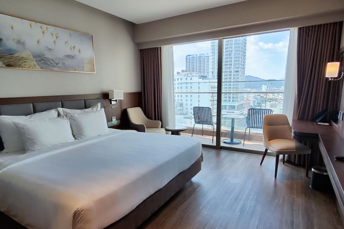Habitación de hotel con una cama grande, un escritorio, dos sillas y vistas a un paisaje urbano a través de amplios ventanales.