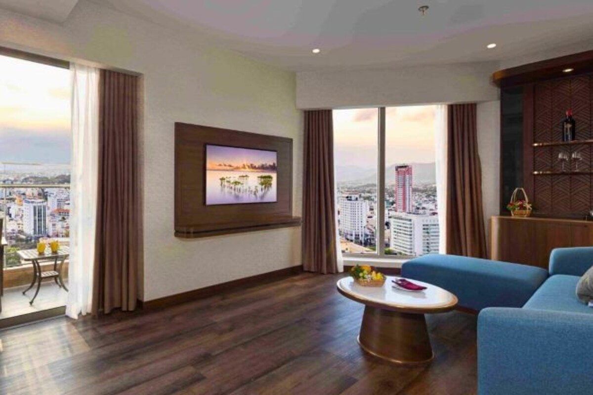 Une chambre d’hôtel moderne dotée d’une grande fenêtre donnant sur la ville, d’un téléviseur à  écran plat fixé au mur, d’un canapé bleu et d’une table basse en bois.