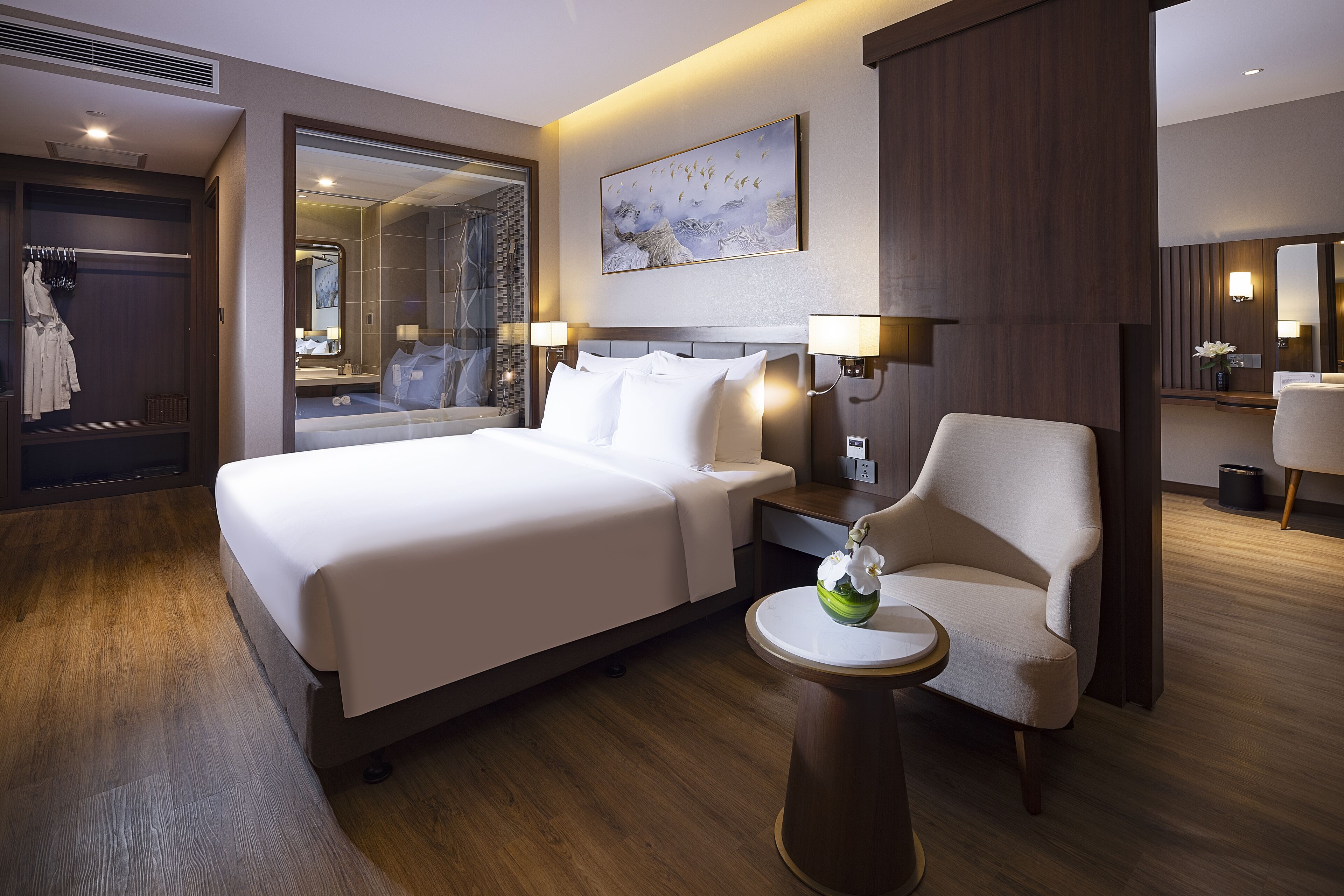 A hotel room with a large bed, a chair, a small table with a flower, and a painting on the wall.