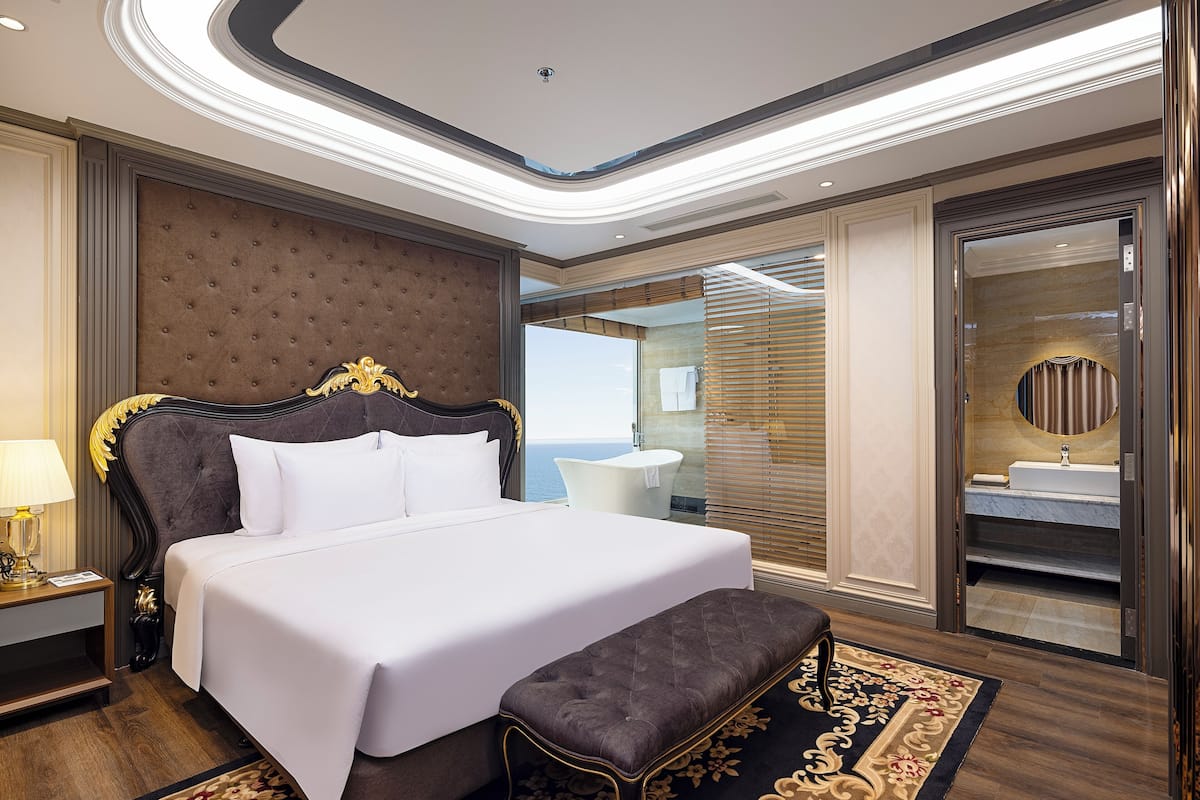 Un dormitorio espacioso con una cama grande, una banca para sentarse, un baño con lavamanos y espejo, y vistas al mar.