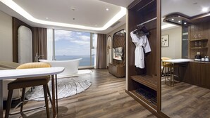 Quarto de hotel moderno com uma janela grande, uma banheira branca, área de estar com mesa e um closet espaçoso com espelho e cabideiro.