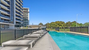 Un área de piscina moderna con muelle de madera, sillas de descanso y piscina de color azul cristalino.